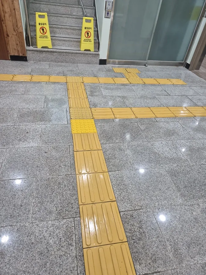계단 앞 출입통제 표지판과 점자유도블록이 교차하는 광택 바닥면